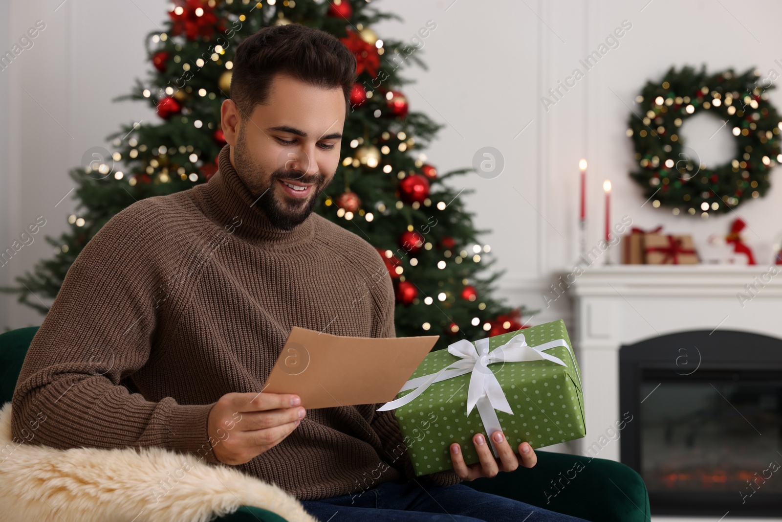 Photo of Happy young man with Christmas gift reading greeting card at home