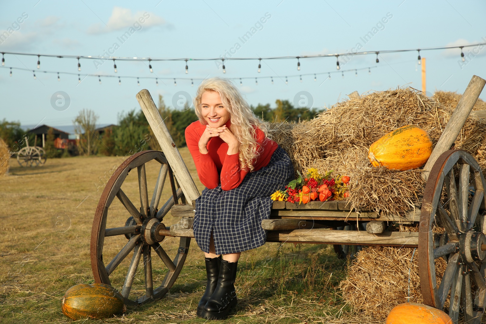 Photo of Beautiful woman with bouquet sitting on wooden cart with pumpkins and hay in field. Autumn season