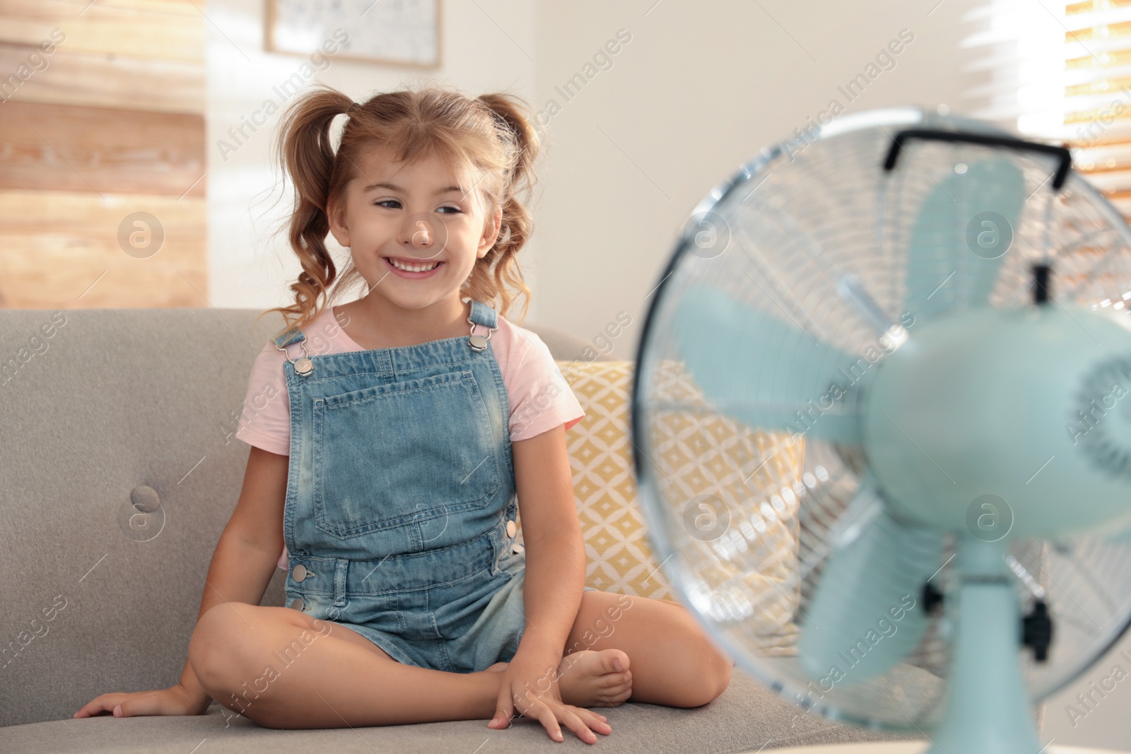 Photo of Little girl enjoying air flow from fan on sofa in living room. Summer heat