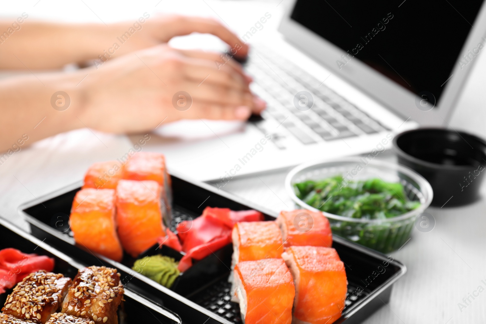 Photo of Boxes with different sushi rolls and blurred woman using laptop on background. Food delivery