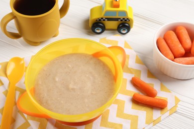 Photo of Baby food. Puree in bowl, small carrots, toy and drink on white wooden table