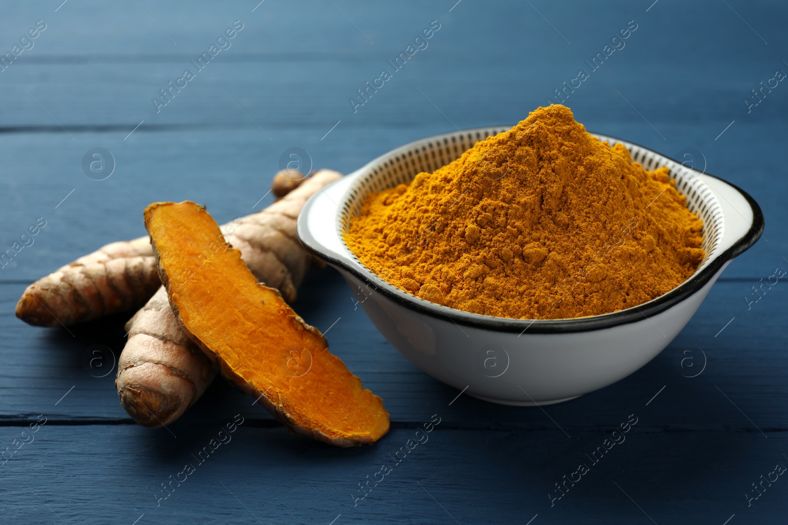Photo of Bowl with turmeric powder and raw roots on blue wooden table, closeup