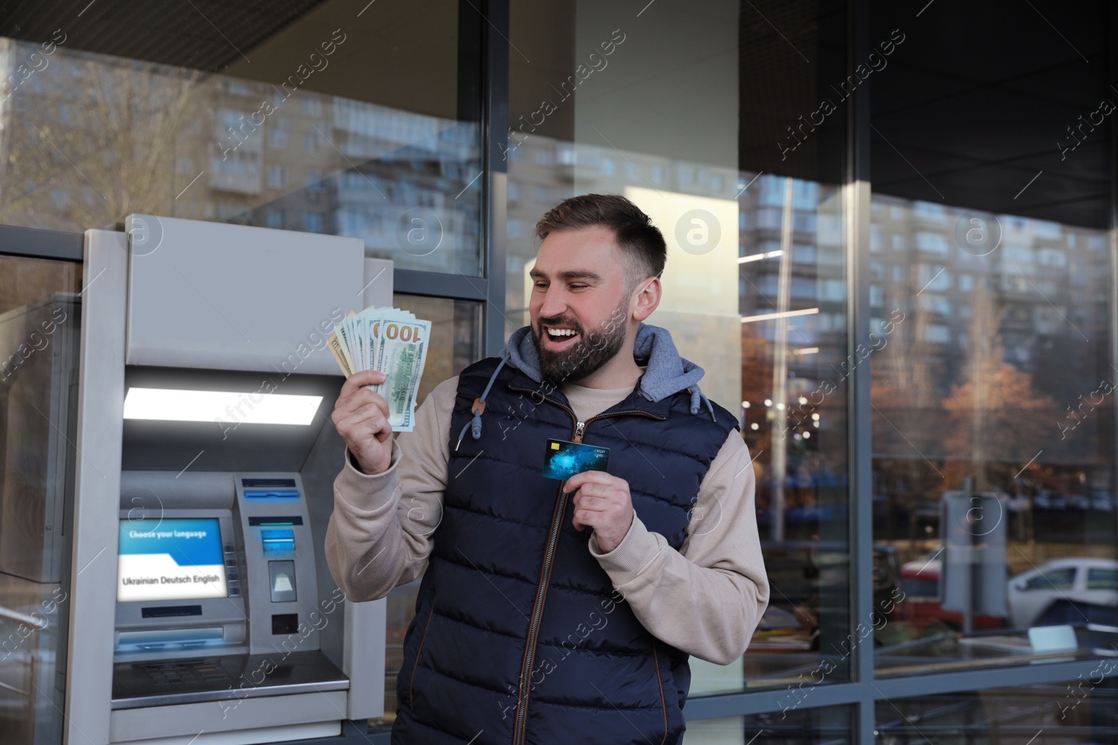 Image of Excited man with credit card and money near cash machine outdoors