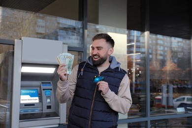 Excited man with credit card and money near cash machine outdoors