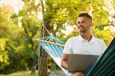 Photo of Young man with laptop in comfortable hammock at green garden