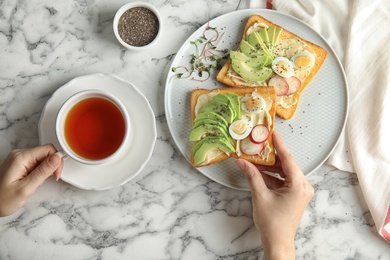 Photo of Woman eating toasts with avocado, quail egg and chia seeds at table, top view