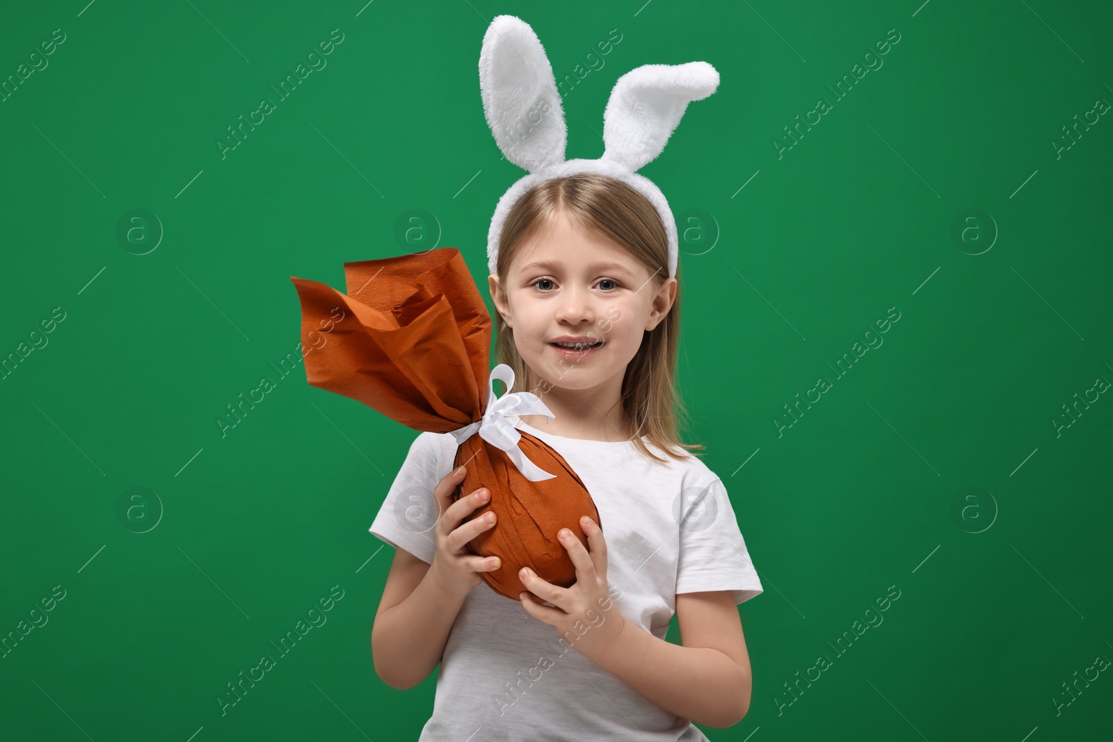 Photo of Easter celebration. Cute girl with bunny ears holding wrapped gift on green background