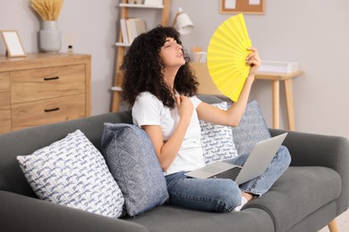 Photo of Young woman with laptop waving yellow hand fan to cool herself on sofa at home