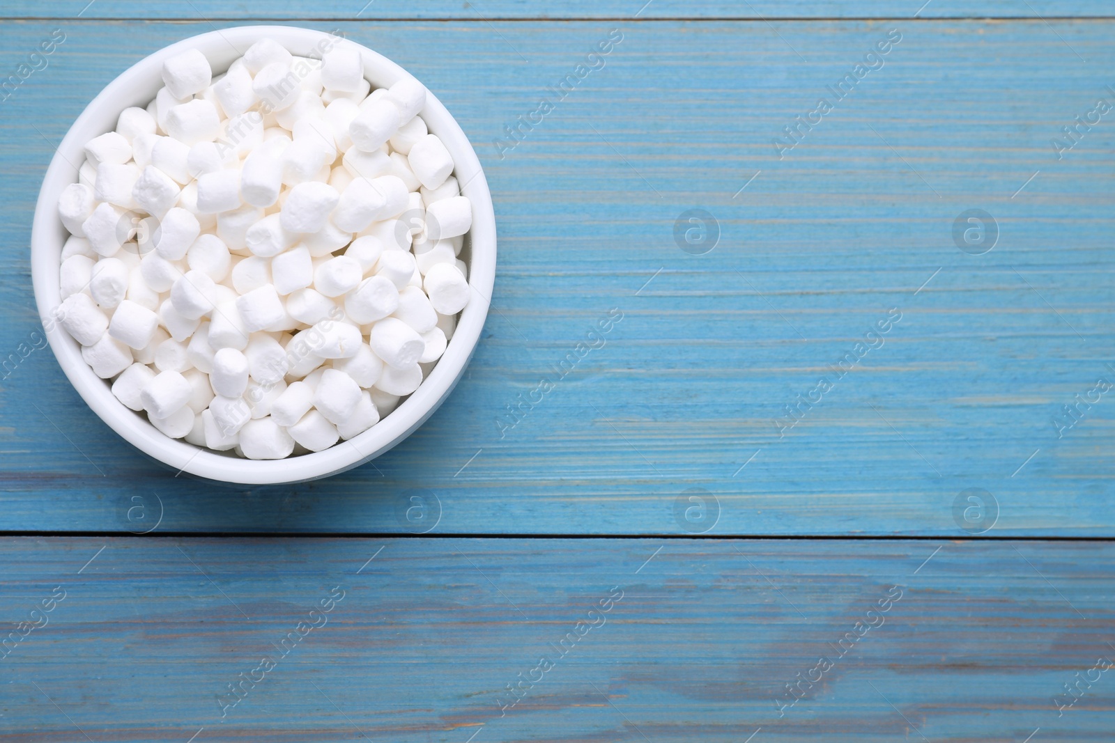 Photo of Bowl with delicious marshmallows on light blue wooden table, top view. Space for text