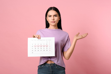 Photo of Young woman holding calendar with marked menstrual cycle days on pink background