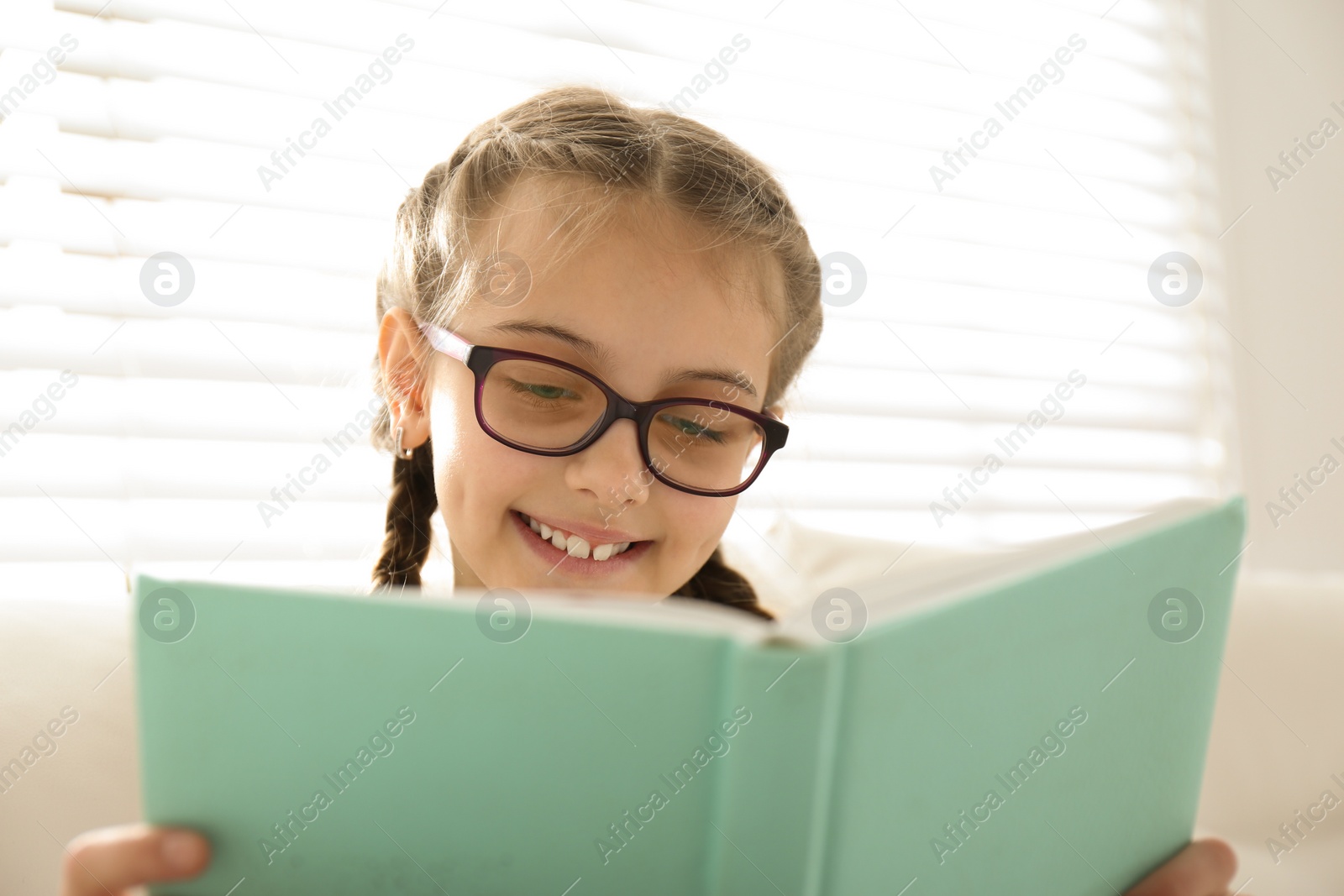 Photo of Cute little girl reading book near window at home