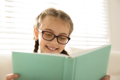 Photo of Cute little girl reading book near window at home