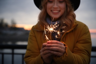Woman in warm clothes holding burning sparkler outdoors, focus on hands