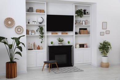 Stylish room interior with beautiful fireplace, TV set and shelves with decor and houseplants