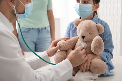 Photo of Pediatrician pretending to examine little girl's bear at hospital. Wearing protective masks
