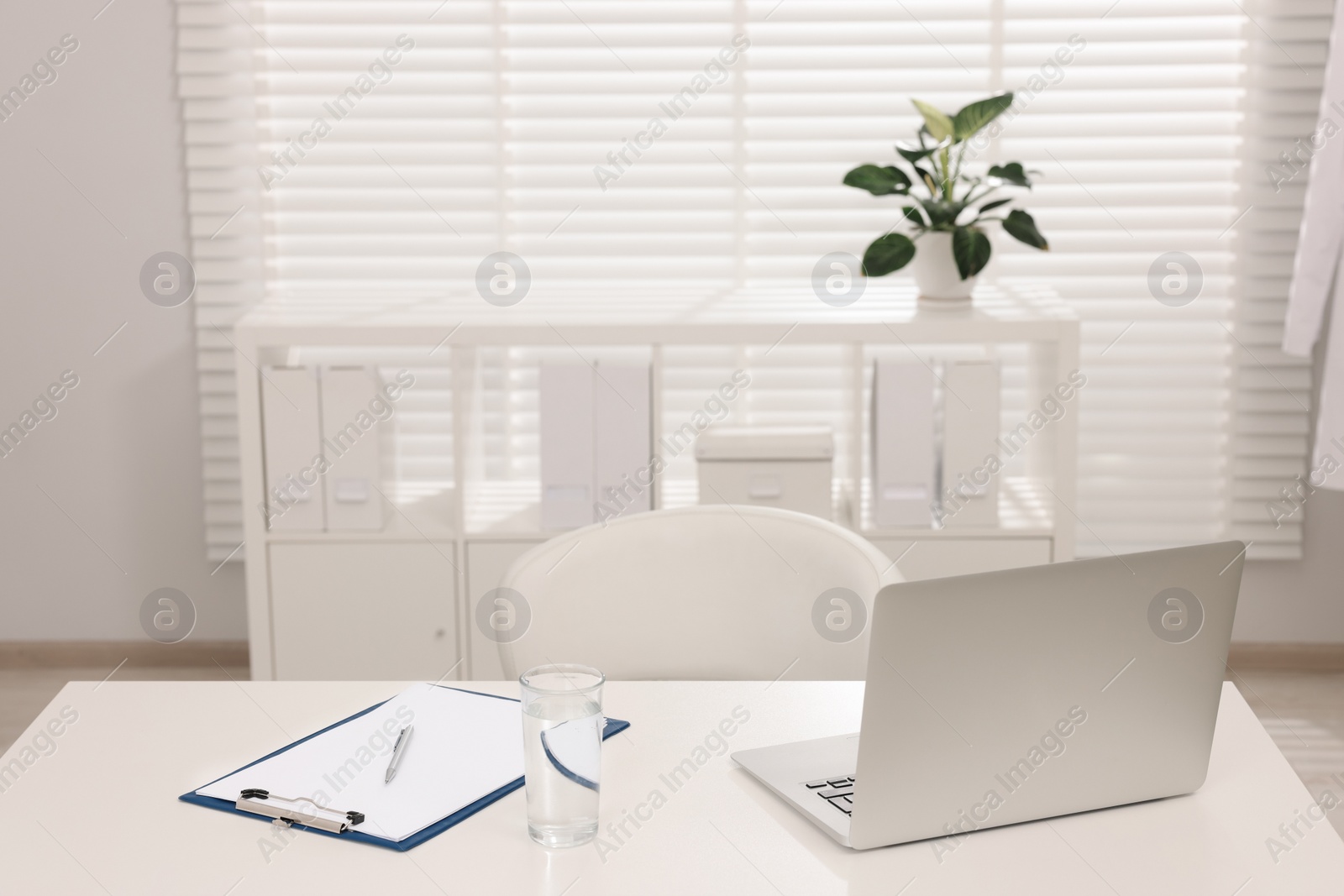 Photo of Doctor's workplace. Laptop, clipboard and glass of water on white table in hospital