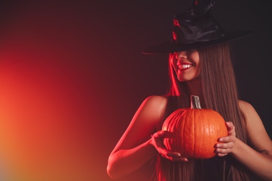 Photo of Young woman wearing witch costume with pumpkin on dark background, space for text. Halloween party