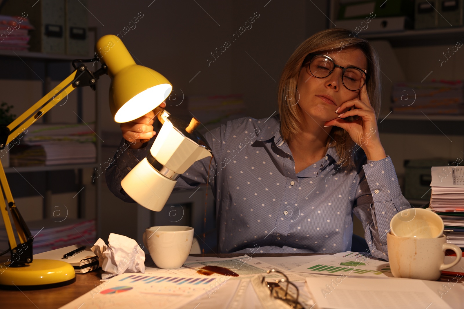 Photo of Overwhelmed woman pouring coffee onto documents while snoozing at workplace in office at night