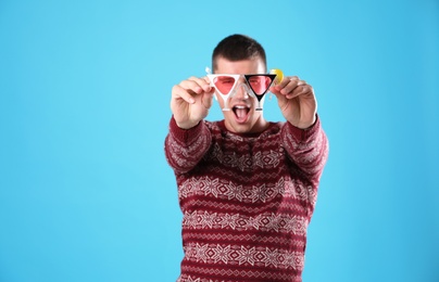 Emotional young man in Christmas sweater and party glasses on light blue background