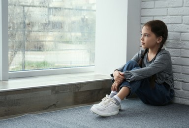 Photo of Sad little girl sitting on floor near window indoors, space for text