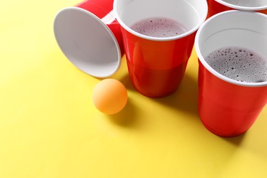Plastic cups and ball on yellow background, space for text. Beer pong game