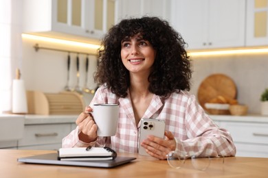 Beautiful young woman in stylish pyjama with smartphone and cup of drink at wooden table in kitchen