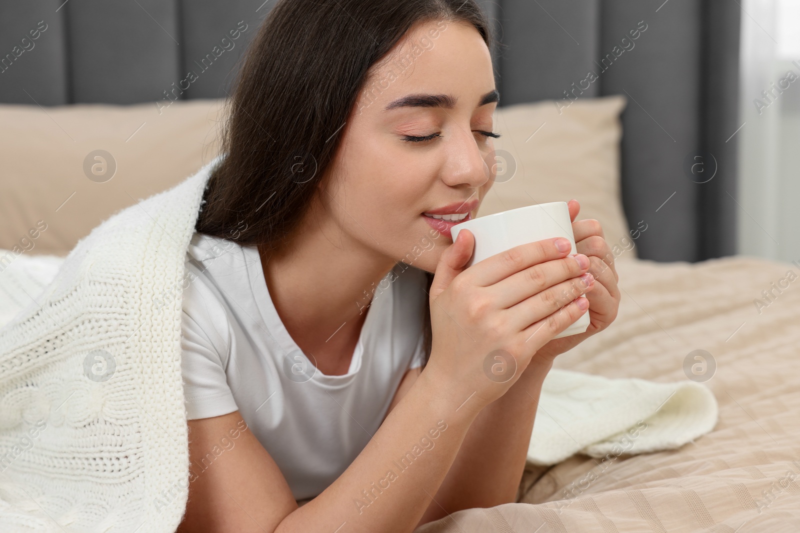 Photo of Beautiful young woman under plaid drinking beverage from white ceramic mug on bed