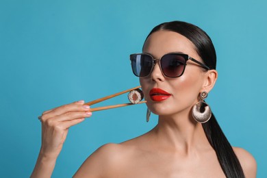 Photo of Attractive woman in fashionable sunglasses holding chopsticks with sushi against light blue background