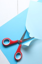 Photo of Red scissors cutting light blue paper on white wooden background, top view