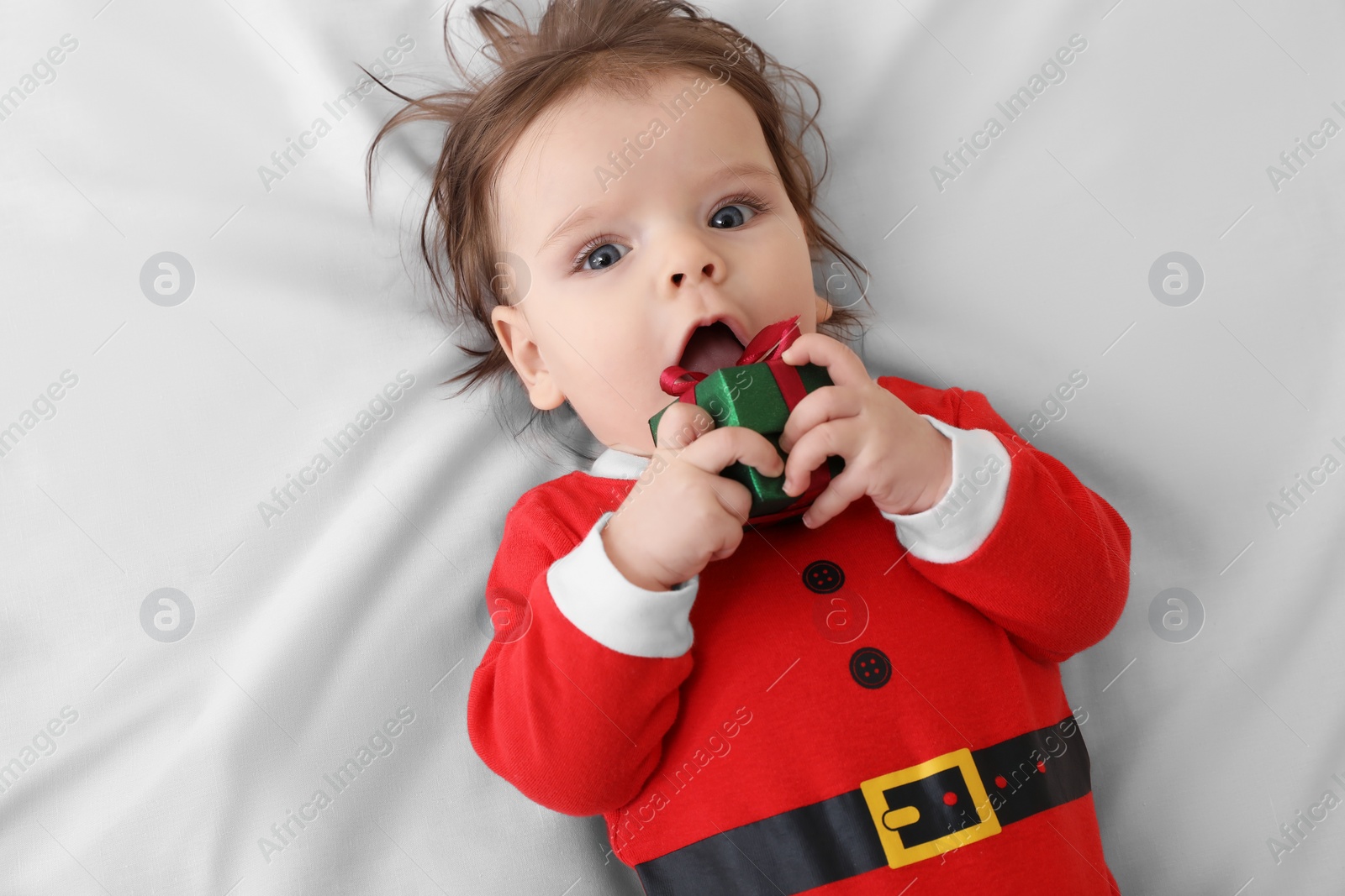 Photo of Cute baby wearing festive Christmas costume with gift box on white bedsheet, top view