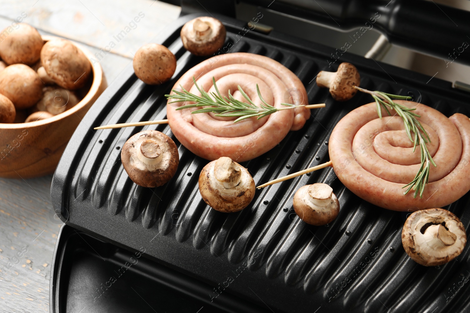 Photo of Electric grill with homemade sausages, rosemary and mushrooms on rustic wooden table, closeup