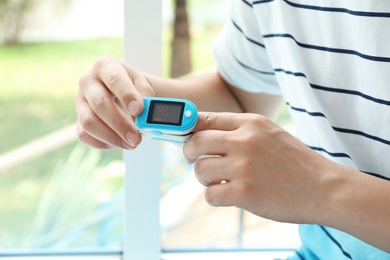 Young man checking pulse with blood pressure monitor on finger against blurred background, closeup