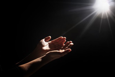 Image of Christian woman stretching hands towards holy light in darkness, closeup. Prayer and belief