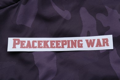 Photo of Paper card with phrase Peacekeeping War on color fabric, top view