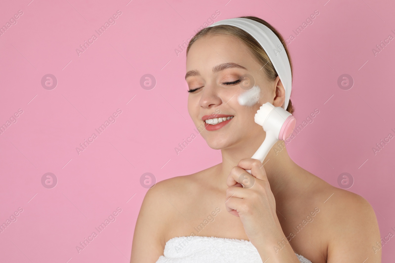 Photo of Young woman washing face with brush and cleansing foam on pink background. Cosmetic products