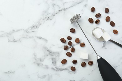 Photo of Black milk frother wand, sugar cubes and coffee beans on white marble table, flat lay. Space for text