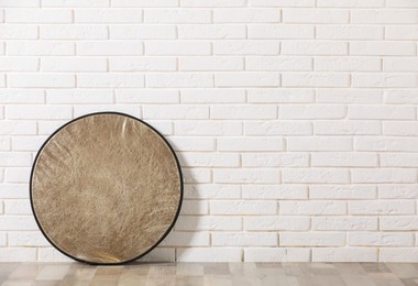 Photo of Professional golden reflector near white brick wall in room, space for text. Photography equipment