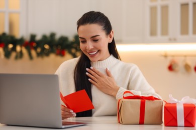 Photo of Celebrating Christmas online with exchanged by mail presents. Smiling woman reading greeting card and gifts during video call at home