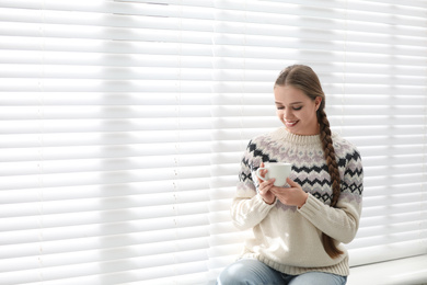 Young woman with hot drink near window at home