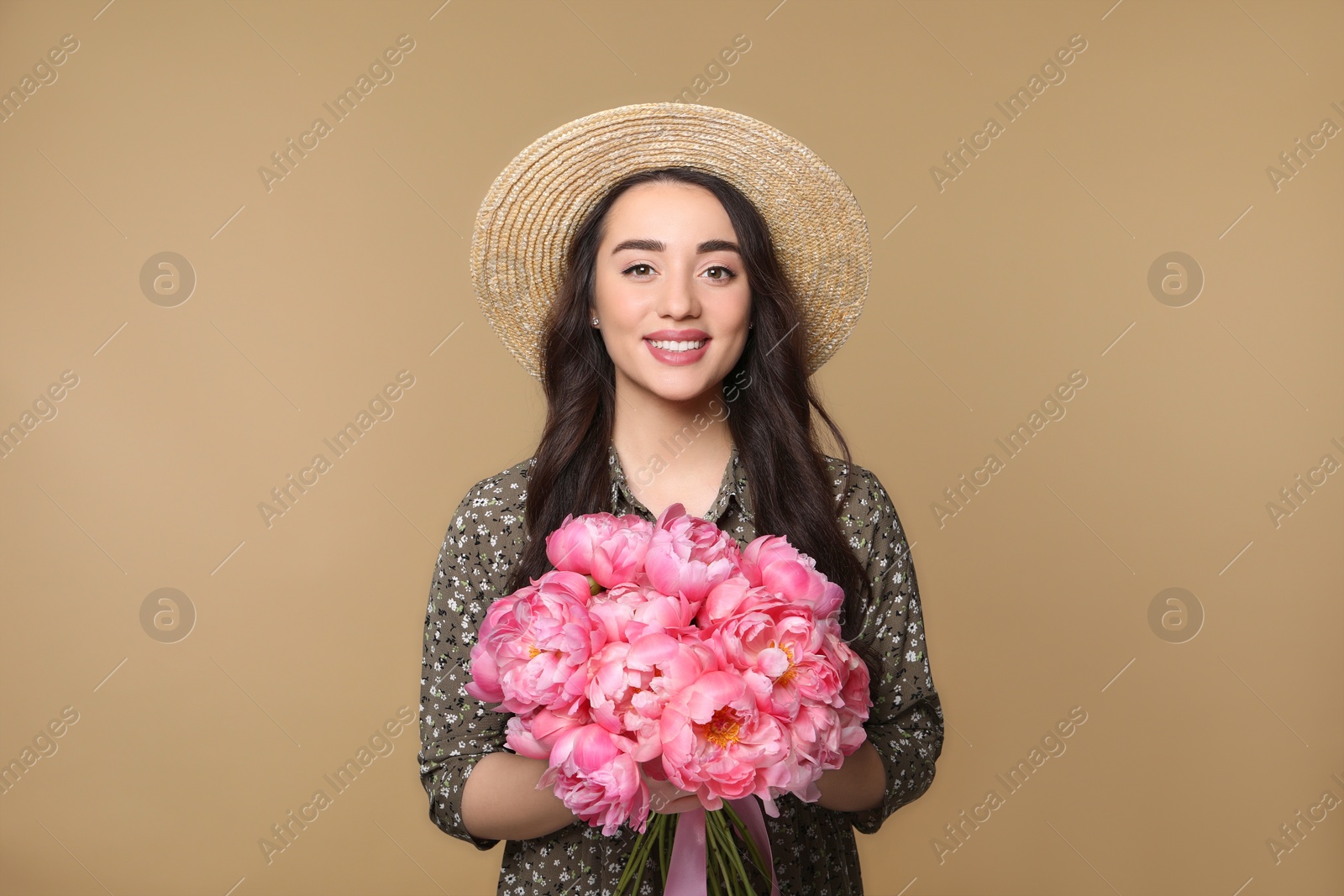 Photo of Beautiful young woman in straw hat with bouquet of pink peonies against light brown background