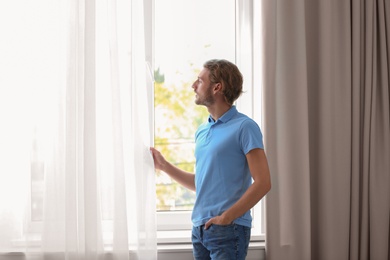 Photo of Young man standing near window with open curtains at home
