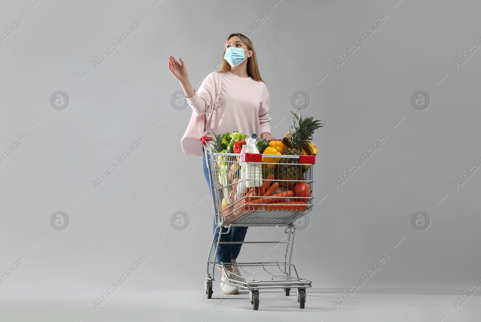 Photo of Young woman in medical mask with shopping cart full of groceries on light grey background