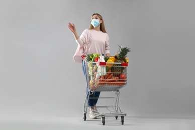 Young woman in medical mask with shopping cart full of groceries on light grey background