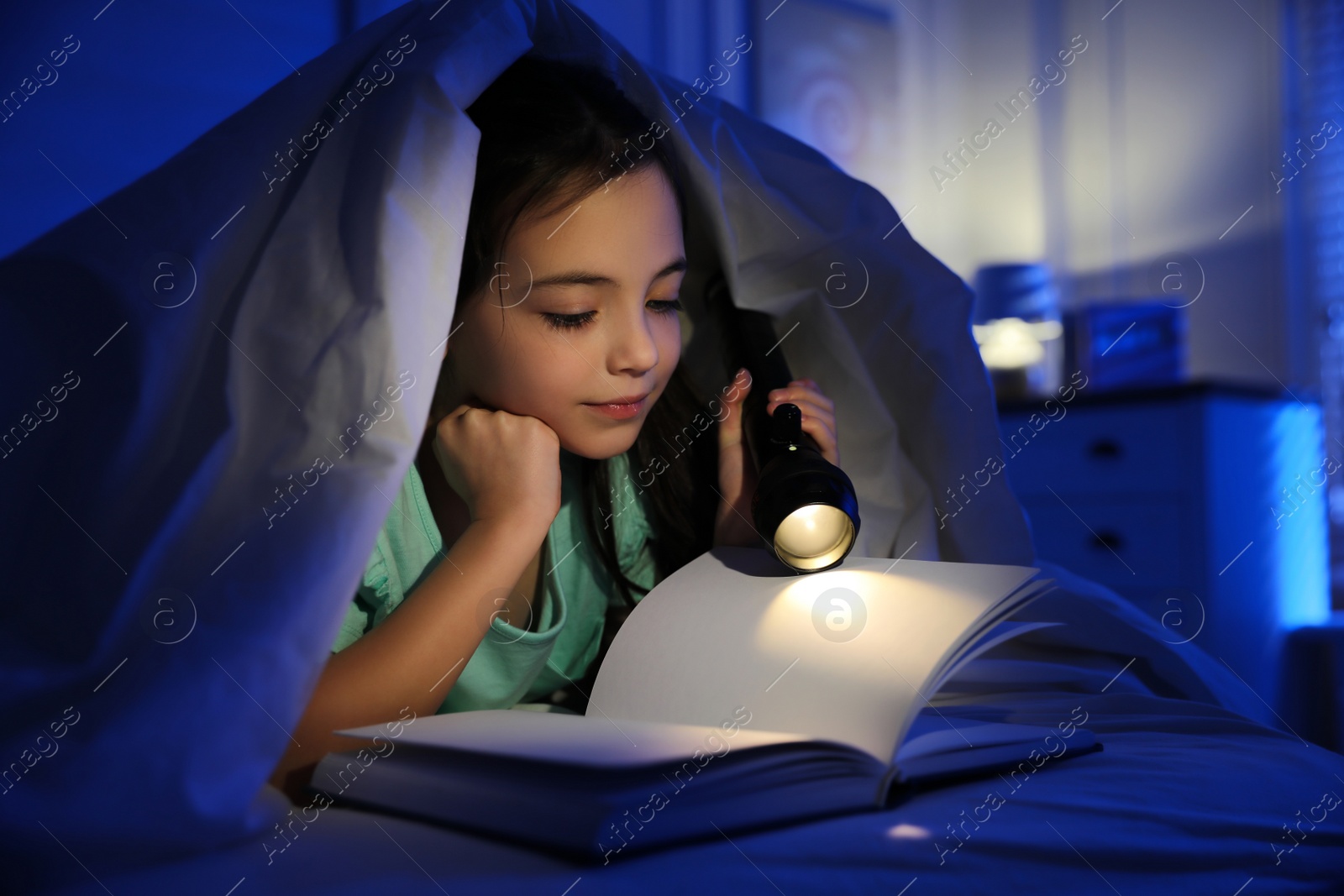 Photo of Little girl with flashlight reading fairy tale in dark bedroom