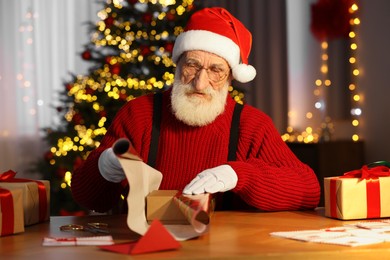 Photo of Santa Claus wrapping gift at his workplace in room decorated for Christmas