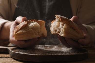 Photo of Man breaking loaf of fresh bread at wooden table, closeup