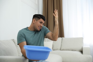 Photo of Emotional man calling roof repair service while collecting leaking water from ceiling in living room
