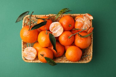 Photo of Fresh ripe tangerines with leaves in wicker basket on green background, top view
