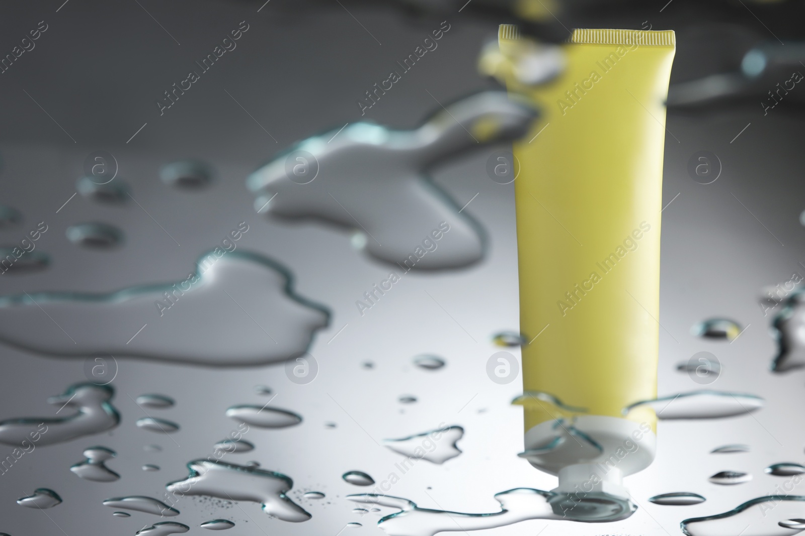 Photo of Moisturizing cream in tube on glass with water drops against grey background, low angle view. Space for text