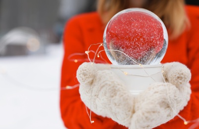 Photo of Woman with knitted mittens holding snow globe and Christmas lights outdoors, closeup. Space for text